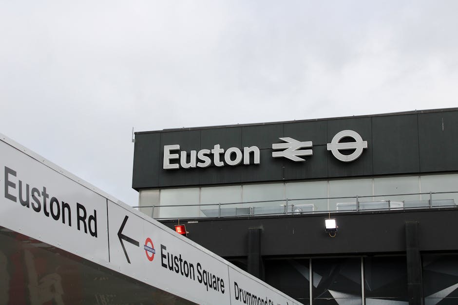 Photograph of the exterior of Euston station showing the station's sign with the London Underground symbol, the Rail Delivery Group logo, and the station name 'Euston' prominently displayed on the dark-colored building. In the foreground, a white directional sign indicates the way to Euston Road and Euston Square, with black lettering and a red and blue London Underground roundel. The scene is captured during daytime with an overcast sky, and the image includes a glimpse of the station's entrance area, with minimal visible movement or activity. This setting relates to the transportation infrastructure used during house relocations or furniture transport activities involved in moving services, as provided by companies like Man with Van Coulsdon.