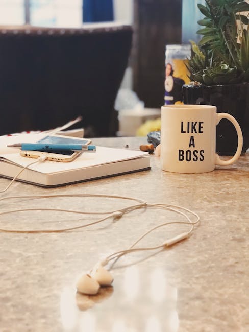 Close-up view of a cluttered indoor workspace with a focus on a beige coffee mug that has the phrase 'LIKE A BOSS' printed in black, positioned on a wooden table. Surrounding the mug are various office items including a closed white laptop with a white charging cable connected, a small stack of blue and white notebooks or folders, and a partially visible potted plant with green foliage in a black container. In the background, there are additional objects such as a plastic water bottle, blurred domestic items, and furniture suggesting a home or office environment. The scene includes natural lighting with some shadowing, and the overall ambiance indicates a casual work setting, possibly during a break or planning stage related to home moves or packing activities.