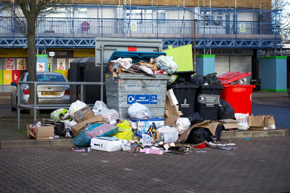 An outdoor scene showing a large pile of waste and refuse bins situated on a paved area in front of a retail premises, with commercial shops and signage visible in the background. The waste includes overflowing black, grey, and red bins, with their lids open, revealing mixed household rubbish, shredded paper, cardboard boxes, plastic packaging, and crumpled bags. Additional discarded items such as flattened cardboard boxes, paper bags, and plastic wrapping are scattered on the pavement around the bins, some partially on a small curb. A metal railing separating the waste area from the parking space can be seen, along with a silver hatchback car parked nearby. The image captures the clutter and mess typical of a collection point for bulky waste disposal, relevant to professional waste removal and clearance services, with the environment under natural daylight.