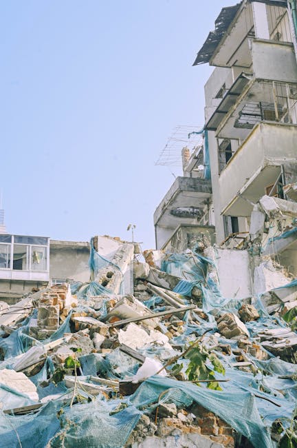 Image showing a pile of mixed debris including bricks, broken concrete, and scattered blue tarpaulin sheets in front of a collapsed or partially demolished multi-storey building. The building features exposed concrete, metal railings, and damaged balconies with some materials hanging or leaning over the rubble. In the background, parts of the structure are still intact, with visible windows and balconies on the upper floors. The sky above is clear and blue, indicating daytime. This scene illustrates a site potentially requiring clearance services as part of a home or property removal process, with a focus on clutter removal and debris disposal, suitable for professionals such as Man with Van Coulsdon specializing in removals and clearance options.