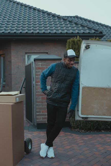 A man wearing a dark cap and jacket is seen partially inside the open driver's side door of a white moving van parked on a paved street. The van's large windshield, side mirror, and headlight are visi