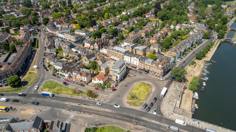 Aerial view of a small town with a mix of residential and commercial buildings, showing narrow streets and a waterfront area with small boats docked along the quay. In the foreground, there is a parki