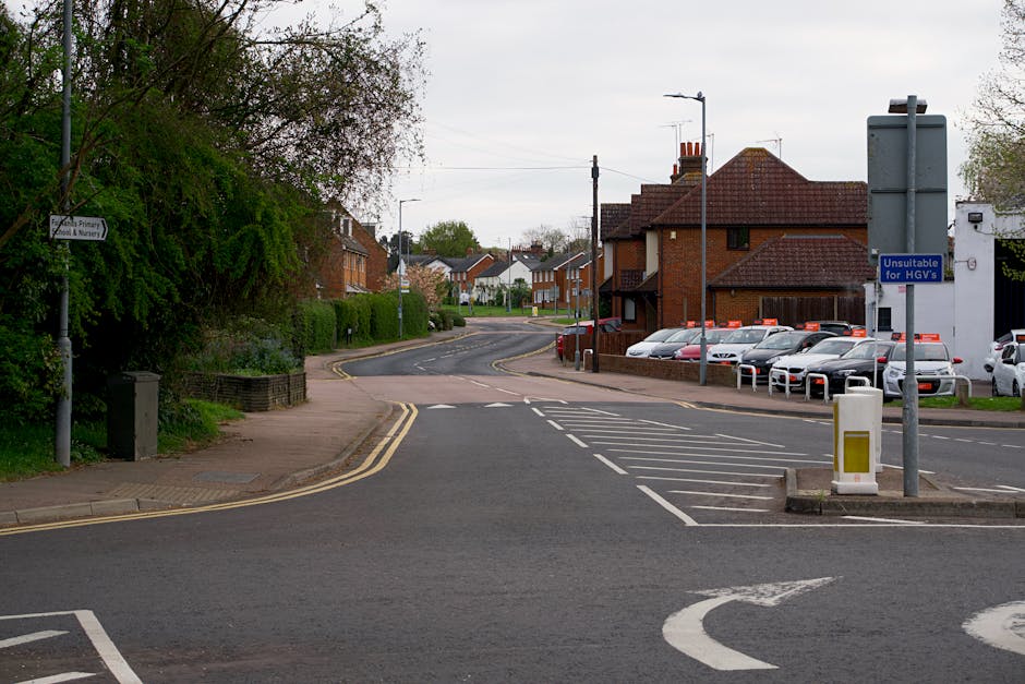 A quiet residential street in Coulsdon with a wide pavement on the left side, bordered by greenery and trees, and a row of parked cars along the curb on the right side. In the background, there are red-brick houses with tiled roofs and a slight curve in the road. A blue sign indicating 'Unsuitable for HGVs' is mounted on a pole near the parking area, which is partially shaded and organized with lines demarcating parking spaces. Streetlights and utility poles are visible along the street, and the scene appears to be during daylight under overcast skies. The environment suggests a typical suburban neighbourhood where house removals may take place, with ample space for loading and unloading furniture or boxes, possibly managed by a local removals company like Man with Van Coulsdon, supporting home relocation and furniture transport services.