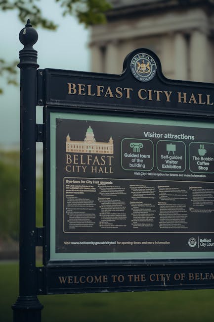A detailed outdoor information board for Belfast City Hall mounted on black metal posts with decorative finials, displaying text and icons about visitor attractions, guided tours, self-guided visits, and the Belfast Coffee Shop. The board features a graphic of Belfast City Hall in gold, green, and white colors, along with the official Belfast City Council emblem at the top. Behind the sign, there are green trees and a cloudy sky, indicating a park or outdoor area. This setting exemplifies a typical location for community notices and may be relevant in the context of local house removals or residential relocation services such as those provided by Man with Van Coulsdon, highlighting the area's amenities and community infrastructure.