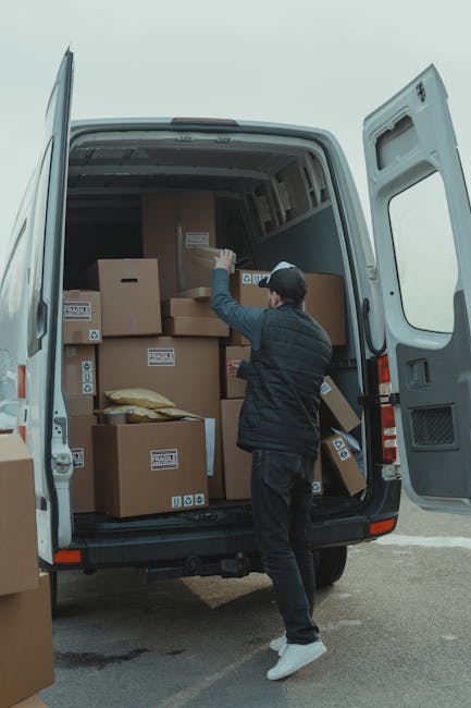 A person wearing a black jacket, dark pants, and a cap is standing at the open rear door of a white moving van, loading or organizing a variety of cardboard boxes of different sizes, some labeled with packing or shipping symbols. The interior of the van is filled with stacked boxes and packing materials, including a yellow padded envelope. The van is parked outside on a paved surface, adjacent to a property, with a clear sky visible in the background. The scene captures the process of home relocation or furniture transport, illustrating the loading stage during a professional removals assignment by Man with Van Coulsdon, supporting efficient packing and moving services on route for a house move in Coulsdon, Croydon.