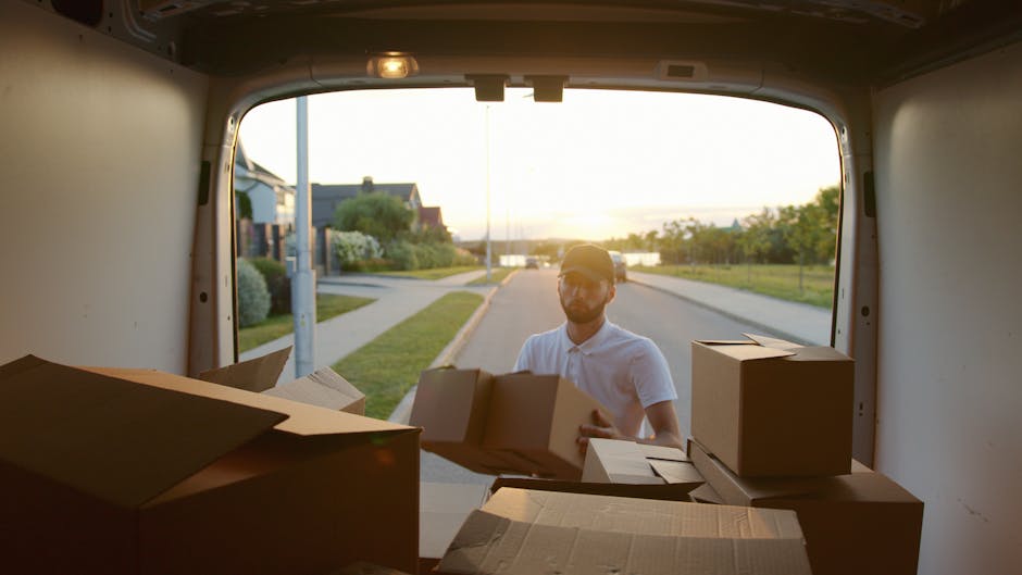 A man is standing outside during sunset, seen through the open rear door of a white moving van with a slightly reflective interior. He is wearing a light-colored baseball cap and a white polo shirt, and is engaged in lifting or placing cardboard boxes into or out of the van. The boxes vary in size, with some fully packed and others partially open, revealing packing materials like paper and bubble wrap inside. The surrounding environment includes a quiet suburban street with well-maintained pavements, residential houses, green lawns, and trees, all bathed in warm, natural light from the setting sun. This scene captures a typical home relocation process involving furniture transport and packing, facilitated by a professional moving service such as Man with Van Coulsdon, with attention to careful handling of household belongings during loading or unloading for a house move in Coulsdon.