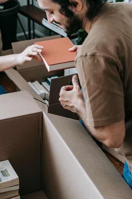 A person wearing a brown t-shirt is seen leaning over a large open cardboard moving box filled with stacked books of various sizes and colours, some with visible text on the spines. The individual is 