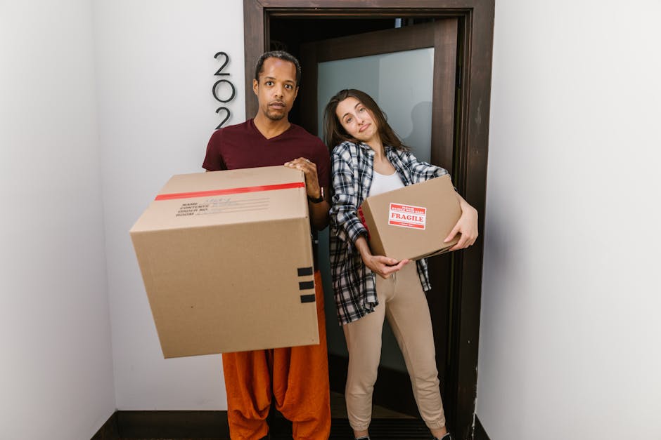 A person wearing a brown t-shirt is seen leaning over a large open cardboard moving box filled with stacked books of various sizes and colours, some with visible text on the spines. The individual is 