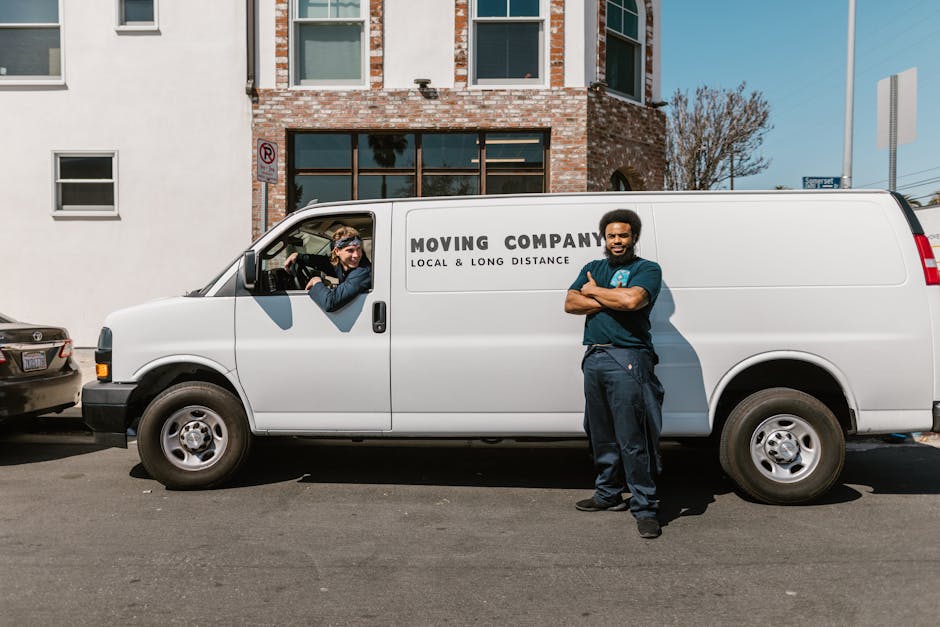 A professional mover from Man with Van Coulsdon poses with arms crossed beside a white moving van labeled 'Moving Company, Local & Long Distance,' parked on the street in front of a residential building with brick and white exterior walls and large windows. The van is positioned parallel to the curb, with the driver’s side door open showing a staff member inside, wearing gloves and glasses, who appears to be preparing or loading household belongings. The scene includes cardboard boxes, wrapped furniture, and plastic packing materials placed near the back of the van, indicating an active home relocation process. Additional parked cars are visible on either side, and the environment is well-lit with natural daylight, suggesting daytime loading or packing during a house move managed by Man with Van Coulsdon, supporting rapid and organized furniture transport and packing services.