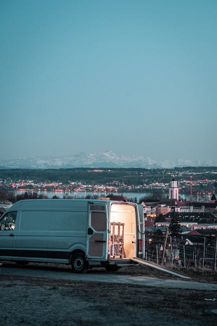 A grey commercial van parked on a paved area in an outdoor setting during twilight, with its rear doors open revealing a wooden chair and packing materials inside, including cardboard boxes wrapped in plastic film. The van is positioned near the edge of the pavement, with a ramp extending to facilitate loading or unloading furniture. In the background, there is a panoramic view of a cityscape with various buildings and industrial structures, and snow-capped mountains further in the distance under a clear evening sky. This scene depicts the loading process involved in home relocation or furniture transport as part of a moving service by Man with Van Coulsdon, supporting swift and efficient house removals.