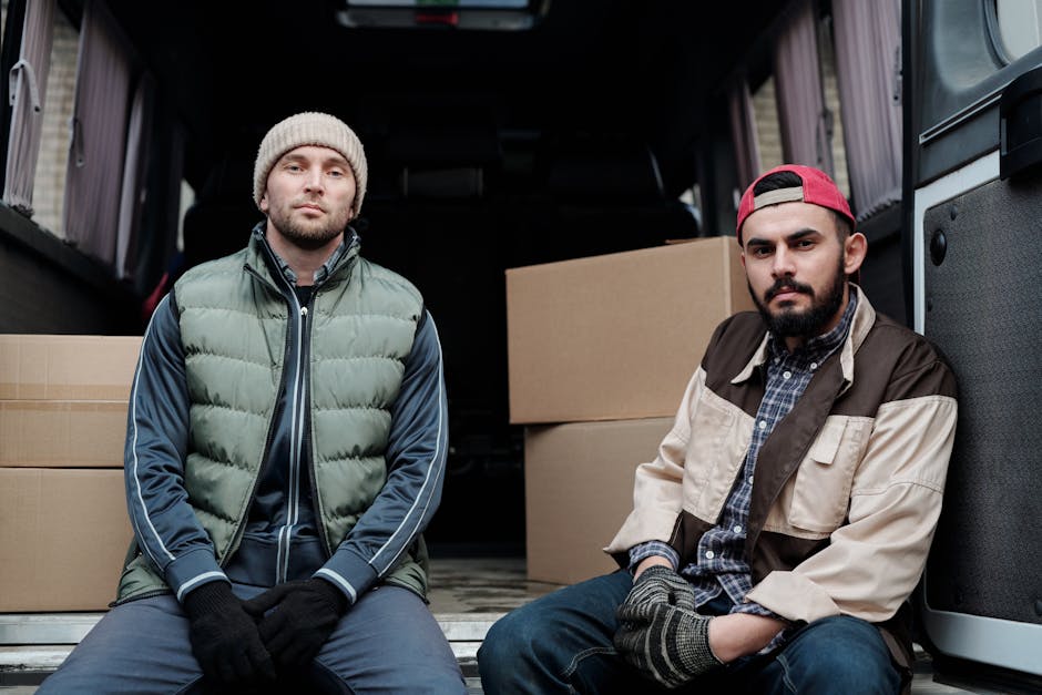 A professional mover from Man with Van Coulsdon poses with arms crossed beside a white moving van labeled 'Moving Company, Local & Long Distance,' parked on the street in front of a residential building with brick and white exterior walls and large windows. The van is positioned parallel to the curb, with the driver’s side door open showing a staff member inside, wearing gloves and glasses, who appears to be preparing or loading household belongings. The scene includes cardboard boxes, wrapped furniture, and plastic packing materials placed near the back of the van, indicating an active home relocation process. Additional parked cars are visible on either side, and the environment is well-lit with natural daylight, suggesting daytime loading or packing during a house move managed by Man with Van Coulsdon, supporting rapid and organized furniture transport and packing services.