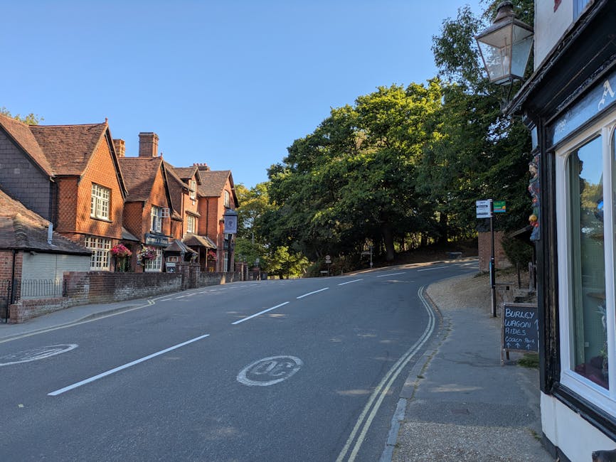 A quiet residential street in Old Coulsdon featuring a gentle upward slope with a double yellow line along the pavement. On the left side, there are traditional brick houses with pitched roofs, some decorated with window flower boxes and small front gardens enclosed by brick walls. To the right, part of a shop or service premises is visible with a large window reflecting the surrounding environment and a blackboard sign advertising burley wagons and rides. A bus stop with a shelter and a bus timetable sign is positioned along the pavement near the shop. The street is lined with mature green trees providing shade and a natural canopy over the scene. The sky is clear and blue, indicating a sunny day. This setting is typical for areas where house removals and furniture transport are carried out, involving careful loading and moving logistics that [COMPANY_NAME] might assist with during home relocation or packing and moving services near Farthing Downs.