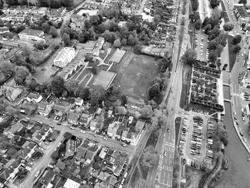 Aerial black-and-white photograph of a residential neighbourhood showing a large open grassy area with sports markings, surrounded by trees and small buildings, adjacent to a busy road with multiple lanes and cars. The road runs alongside a parking lot filled with numerous vehicles, bordered by a row of trees and hedges. To the left, there are residential houses with pitched roofs, small gardens, and driveways. In the foreground, a collection of cardboard boxes, plastic wrapped furniture, and various household items are being loaded into the back of a large van belonging to Man with Van Coulsdon, parked on the pavement near the house. The loading process involves lifting and carrying items towards the vehicle, which is equipped with straps and blankets for safe furniture transport. The scene depicts the indoor-to-outdoor move-in process during house removals, with items inside a property ready to be loaded into the van as part of a home relocation. Natural lighting conditions and a clear spatial arrangement of objects, personnel, and vehicles are visible, emphasizing the logistics involved in packing and moving services near Farthing Downs, Coulsdon.