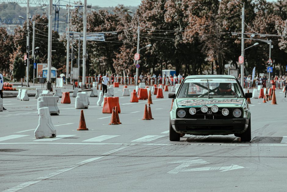 A black vintage-style car with four front-mounted auxiliary lights is parked on a street temporarily closed for an event, with multiple orange and white traffic cones and barriers set up to block access. The vehicle is positioned near a pedestrian crossing, facing towards the camera, with the driver visible through the windshield. In the background, a few pedestrians are walking amongst the cones, near a park with trees and urban infrastructure including streetlights, overhead wires, and traffic signs. The scene suggests a controlled area adapted for a specific purpose, possibly related to the logistical aspects of home relocation or moving services, reflecting organized movement and parking restrictions under local regulations. The image conveys the setting of a street scene where parking suspensions and cones support efficient furniture transport or moving processes, captured in natural daylight with clear visibility.