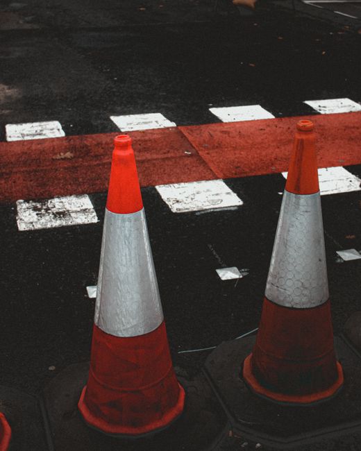 Close-up view of two orange and white traffic cones placed on a paved street surface, with partial white dashed markings and a red textured strip indicating a no-parking or restricted zone. The cones are located near the edge of the road, which appears to be part of a street undergoing moving or transport activity. The background shows a dark asphalt pavement with some scattered debris or dirt. This scene depicts the type of parking suspensions, cones, and safety arrangements often used during house removals and furniture transport in the Coulson area, as managed by Man with Van Coulsdon. The setup suggests preparation for a home relocation or moving process, ensuring vehicle and pedestrian safety during loading or unloading procedures.