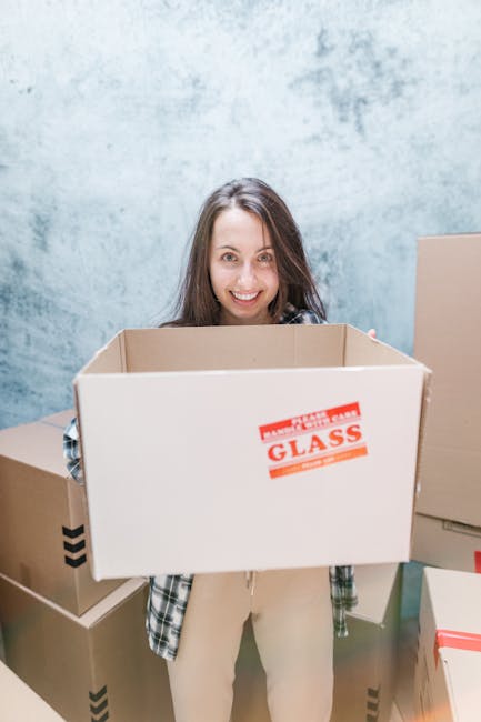 A woman with long brown hair and a friendly expression is standing indoors against a light blue textured wall, holding an open cardboard box in front of her. The box has a red and white label that reads 'Glass' indicating it is designated for fragile items. She is dressed in casual clothing, wearing a plaid shirt layered over a top, and beige trousers. Surrounding her are several other packed cardboard boxes, some sealed with tape and others open, placed on the floor and nearby surfaces. The scene depicts a home relocation or packing process, with the woman preparing items for transport. Visible on the ground are additional boxes stacked and ready for moving, conveying an organized packing effort associated with house removals, facilitated possibly by a professional service such as Man with Van Coulsdon. The environment appears well-lit, indicating a clean, dedicated space for packing activities as part of a furniture transport or moving logistics operation.