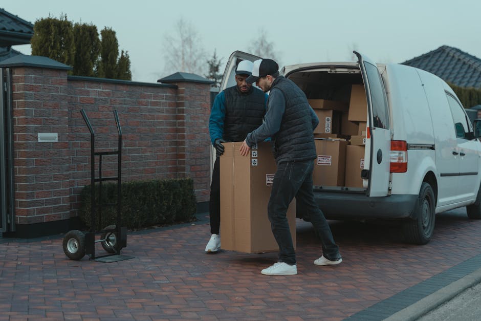 Two men from Man with Van Coulsdon are engaged in the home relocation process, depicted loading or unloading a large cardboard box from a white delivery van parked on a brick driveway. The van's rear 